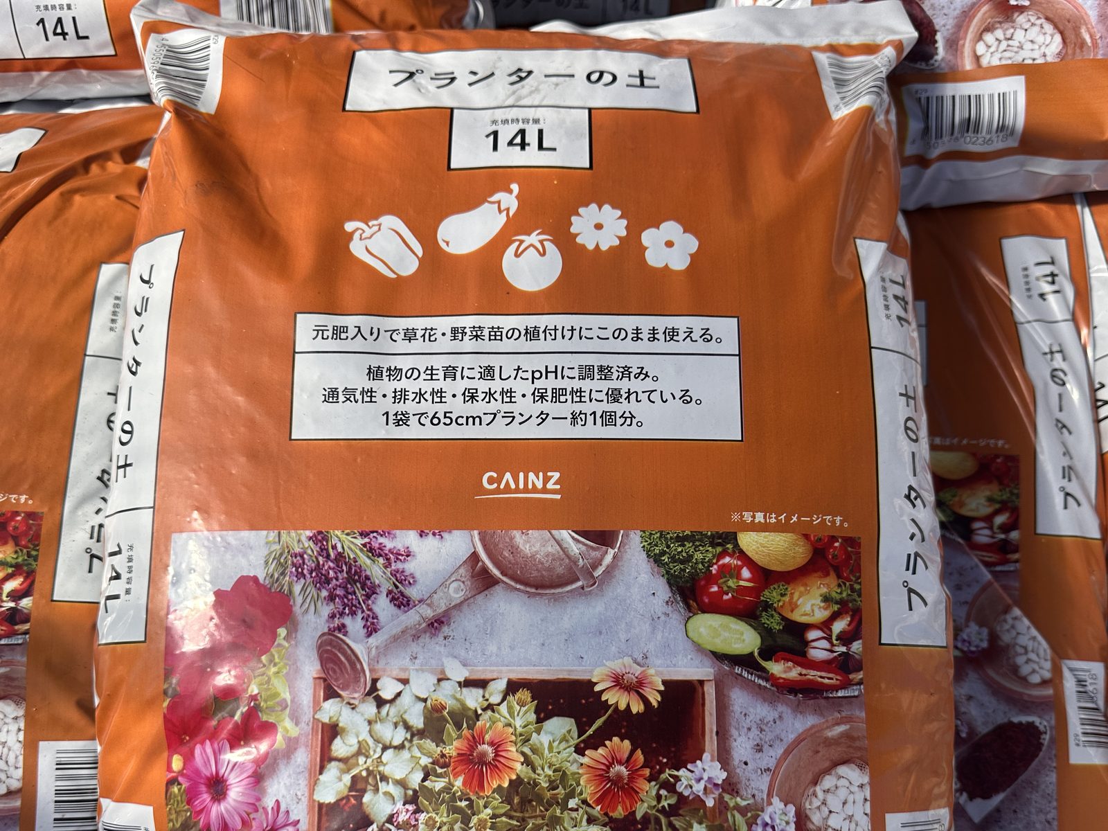 カインズの店頭に積まれた「プランターの土」14L の袋。表に「元肥入りで草花・野菜苗の植付けにこのまま使える。」「植物の生育に適したpHに調整済み。」「1袋で65cmプランター約1個分。」と書いてある