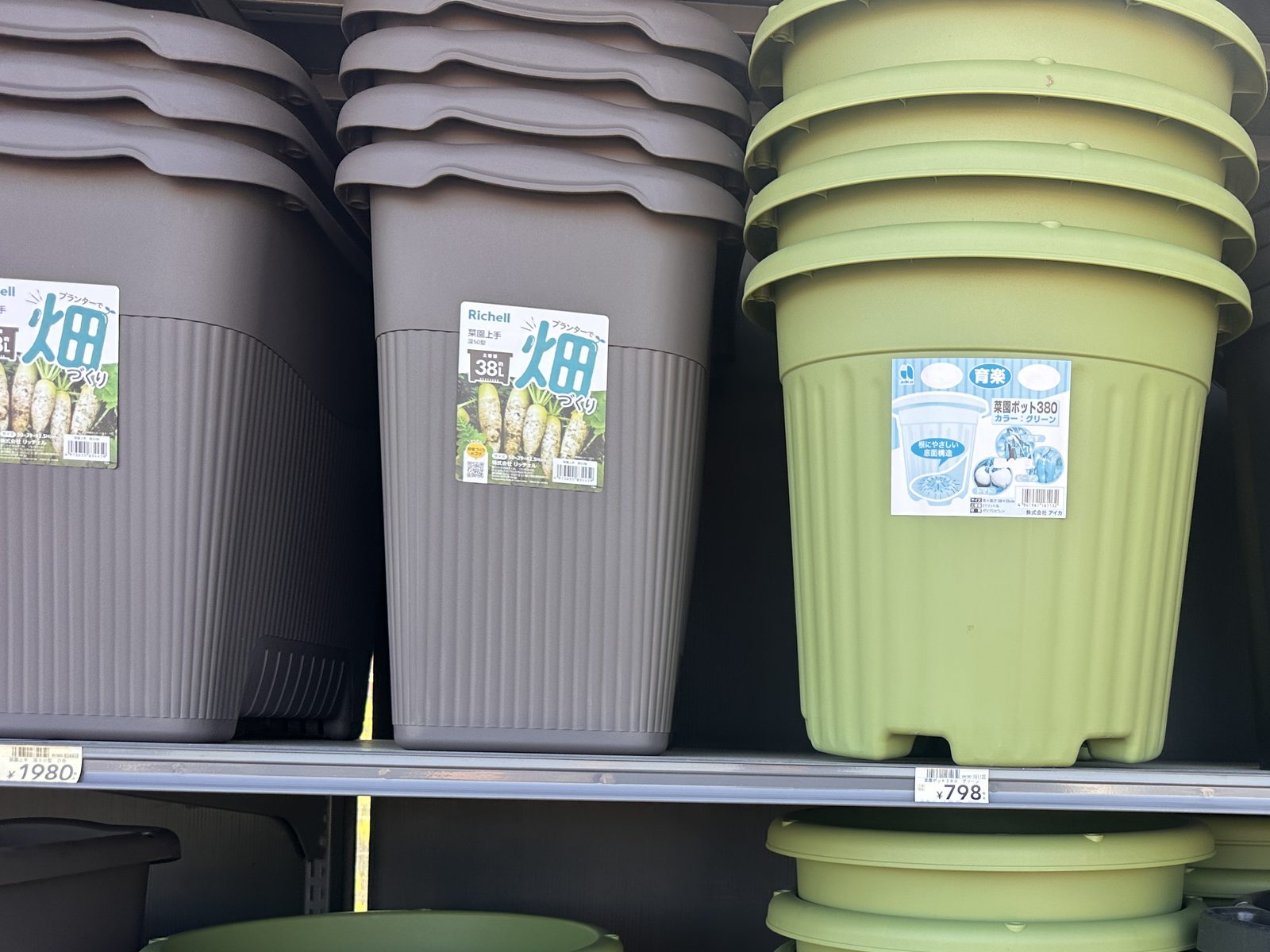 ホームセンターのプランター売り場。茶色い角形の深型プランターに「土容量 約38L」のラベル、隣に緑の丸い鉢が重ねて並ぶ